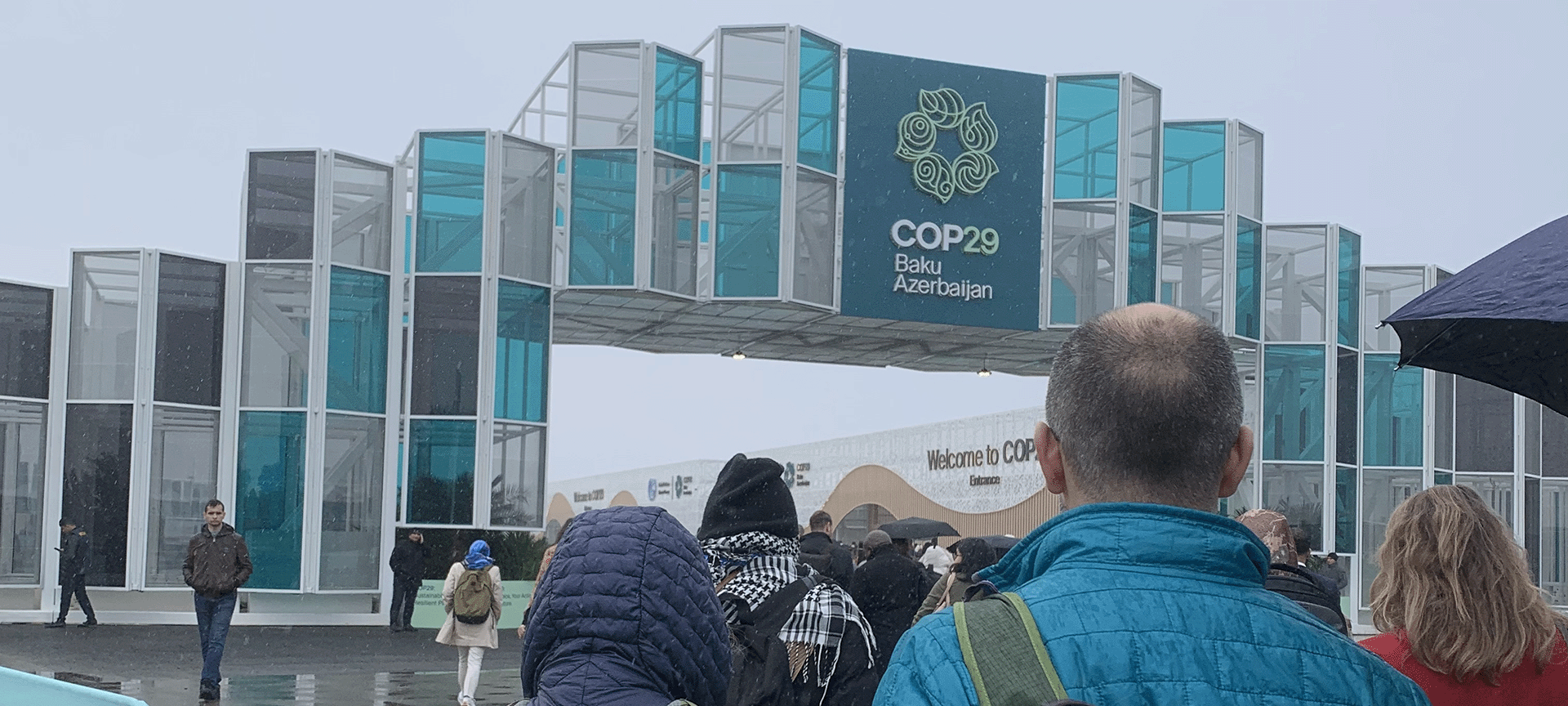 Rain falls on the banner showing the logo of COP29, Baku. People in rain-wear and hoods approach the COP entrance Climate, climate crisis, climate finance, loss and damage