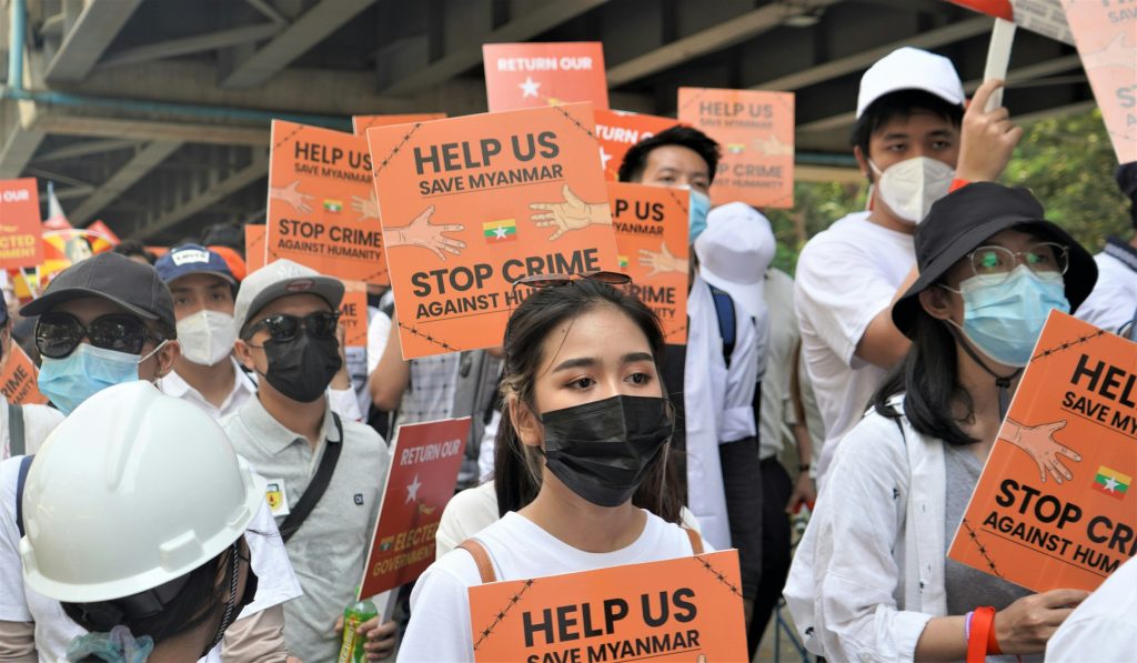 woman in white crew neck t-shirt wearing black sunglasses carries an orange sign 'help us save Myanmar; stop crime against humanity' Myanmar's ongoing crisis